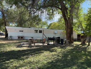 A mobile home with outdoor furniture, a barbecue grill, and a tree in the foreground.