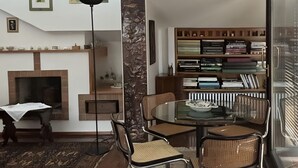 Ein modernes Wohnzimmer mit Kamin, einem Esstisch mit Stühlen, einem Bücherregal und einem großen Fenster.