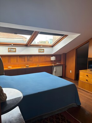 Ein Schlafzimmer mit Dachfenster, Holzvertäfelung, einem Bett mit blauer Bettdecke, einem Nachttisch und einer Kommode.