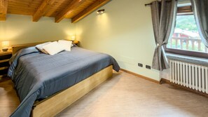 A bedroom with a bed, wooden headboard, bedside lamps, a window with curtains, and a radiator. - Abri Douillet - Cozy Apartment with Private Parking in Saint-Marcel (Saint-Marcel)