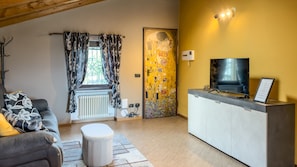 A living room with a sofa, a small table, a radiator, a television on a cabinet, and a door with a colorful mural. - Abri Douillet - Cozy Apartment with Private Parking in Saint-Marcel (Saint-Marcel)