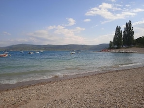 A pebbled beach with calm water, boats, and distant hills.