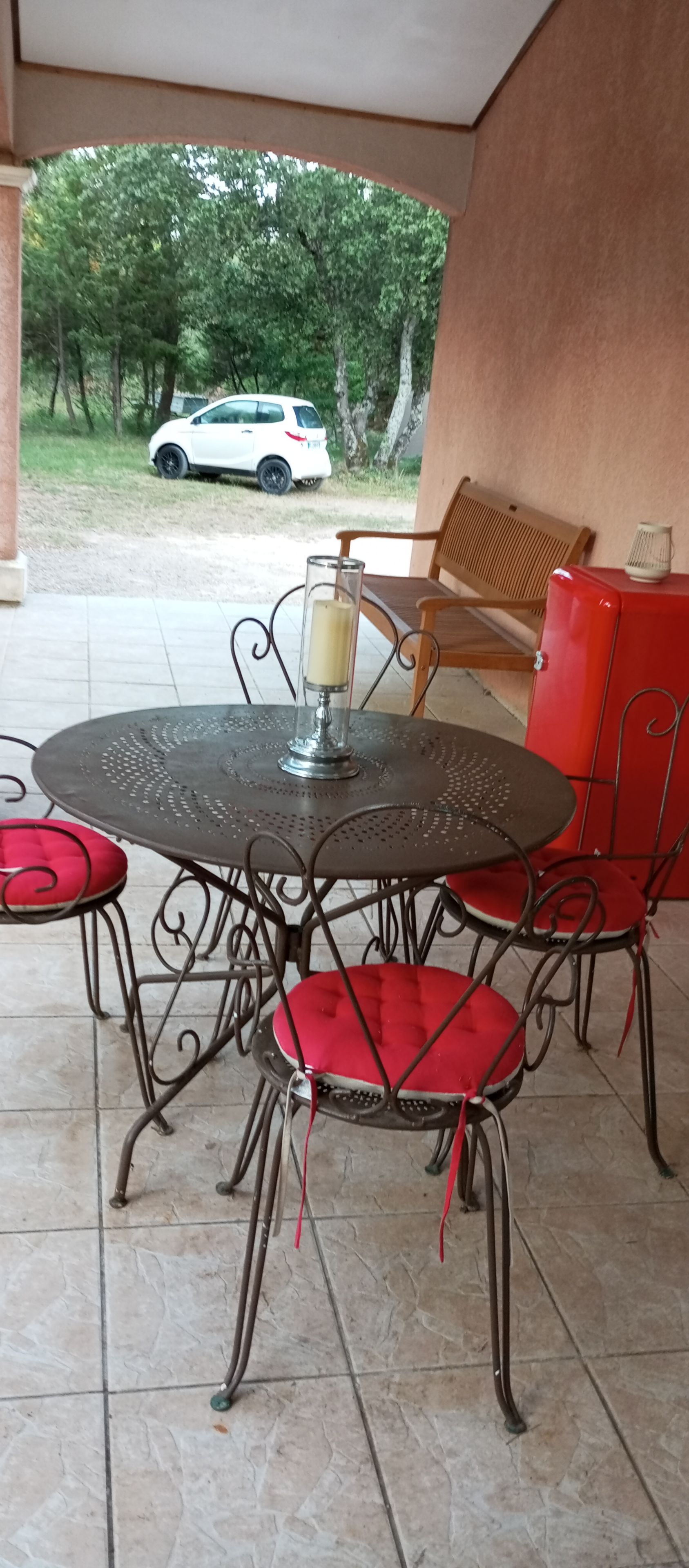 Um pátio externo com uma mesa redonda de metal e cadeiras vermelhas com almofadas, um castiçal de vidro e um carro branco estacionado ao fundo.