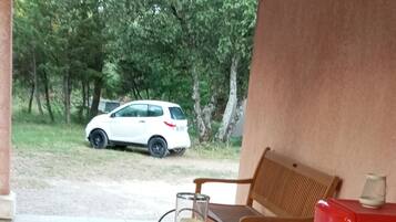 An outdoor patio with a round metal table and red cushioned chairs, a glass candle holder, and a white car parked in the background.