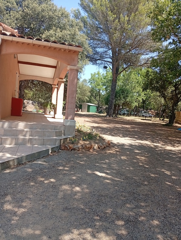A covered outdoor area with steps leading to a building, surrounded by trees and a gravel ground.