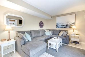 Ein Wohnzimmer mit einem grauen Ecksofa, einem weißen Couchtisch, einem Beistelltisch mit Lampe, einer Wanduhr und einem gerahmten Bild.