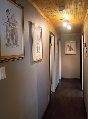 A hallway with framed artwork on the walls, wooden ceiling, and a light fixture.