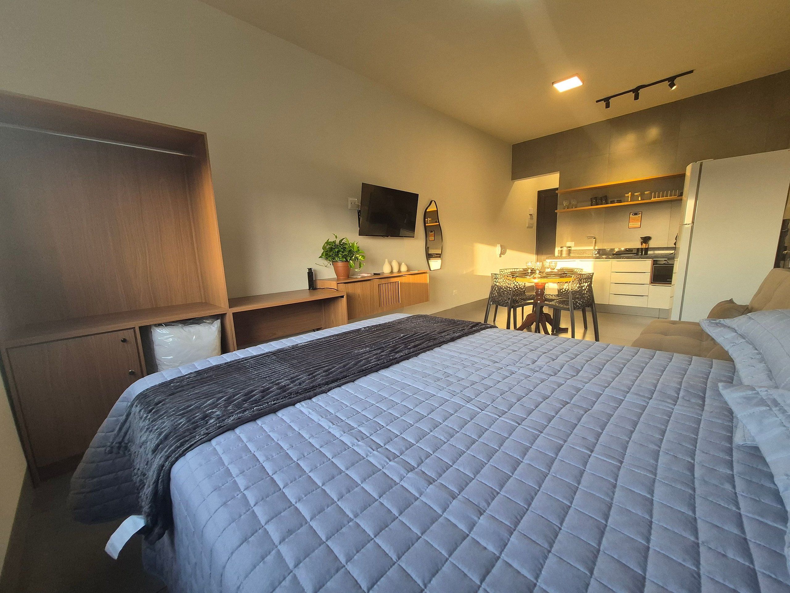 A bedroom with a bed, bedside tables, a desk, a chair, a potted plant, and a kitchen area in the background.