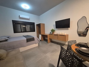Una habitación con cama, escritorio, silla, mesa de comedor, televisor y espejo.