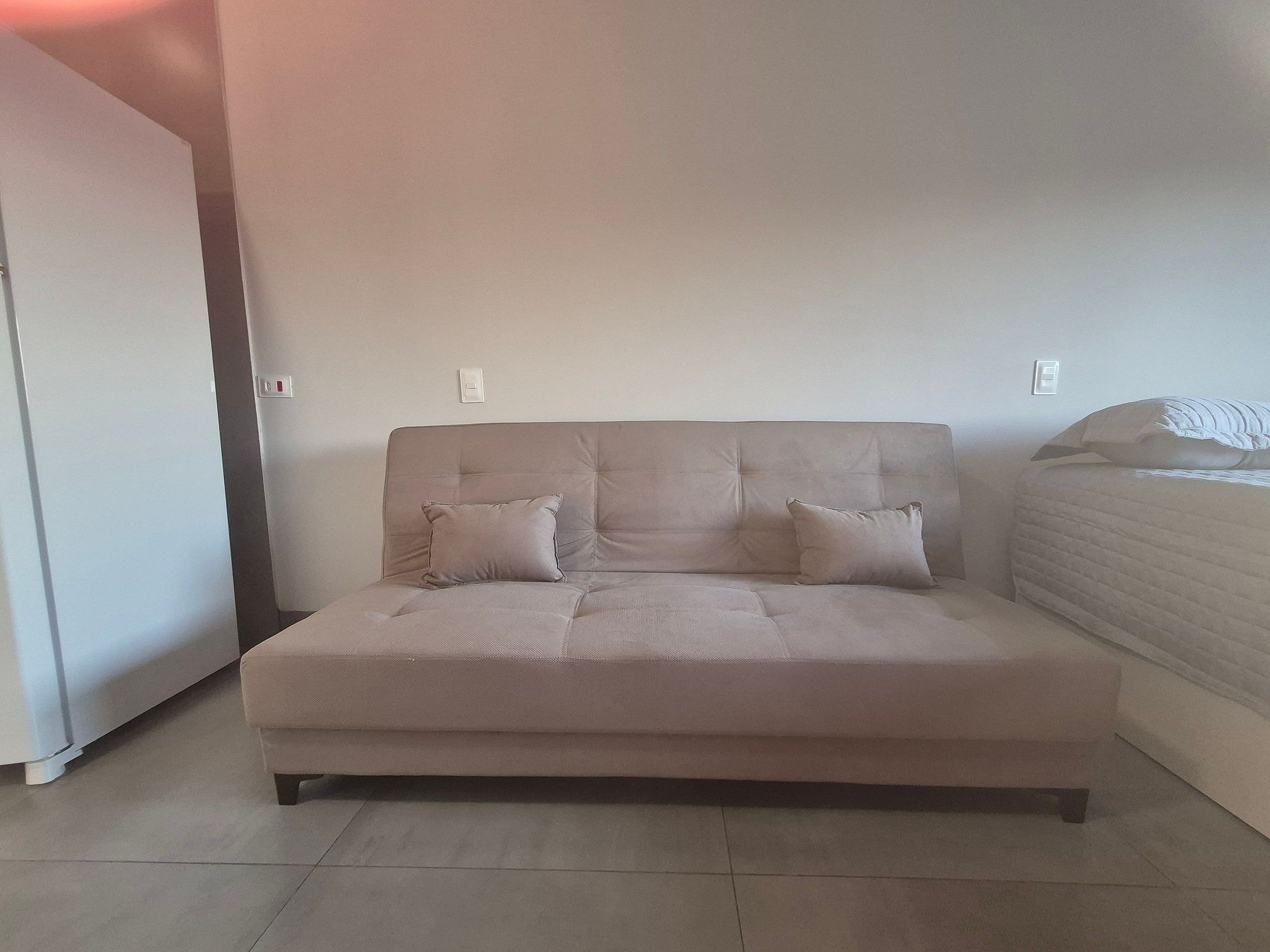 A neutral-toned sofa with two cushions, a tiled floor, and a plain wall with a light switch.