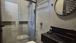 A modern bathroom with a glass-enclosed shower, a white toilet, a black sink, and a mirror with a circular frame. - PP - Maria da Glória - Apto MG01 (Presidente Prudente)