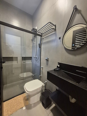 Un baño moderno con ducha envuelta en vidrio, inodoro blanco, lavamanos negro y espejo con marco circular.