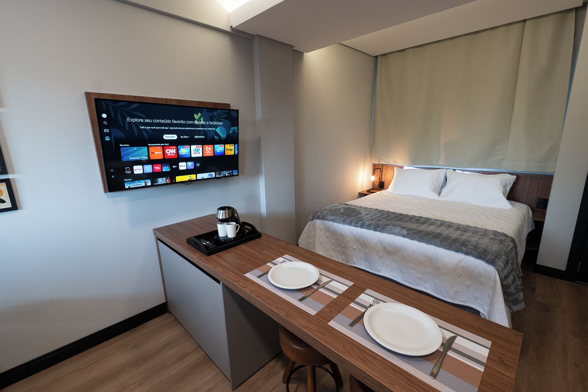 A hotel room with a bed, a desk with a coffee maker, and a TV displaying various channels.