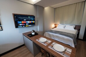 Quarto de hotel com uma cama, uma mesa com cafeteira e uma televisão exibindo vários canais.