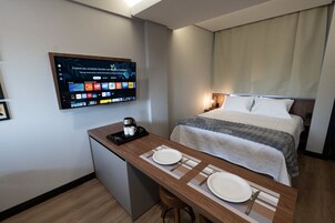 Quarto de hotel com uma cama, uma escrivaninha com cafeteira e uma televisão exibindo diversos canais.