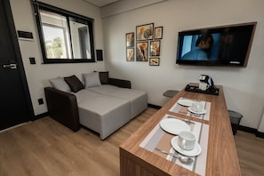 Una sala moderna con un sofá gris, una mesa de comedor de madera dispuesta con platos y tazas, un televisor de pantalla plana montado en la pared y una ventana con persianas.