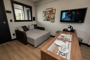 Uma sala de estar moderna com um sofá cinzento, uma mesa de jantar de madeira posta com pratos e chávenas, uma televisão de ecrã plano na parede e uma janela com persianas.