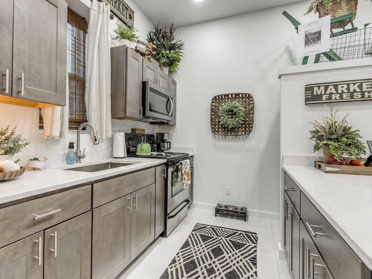 A kitchen with grey cabinets, a microwave, a stove, and a sink. There are plants on the countertops and a rug on the floor.