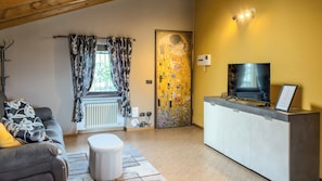A living room with a sofa, a small stool, a radiator, a television, and a door with a floral design. - Abri Douillet (Saint-Marcel)