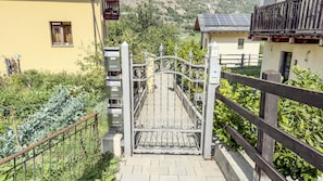 A gated entrance with a metal gate and a wooden fence, leading to a building with a yellow facade and a balcony with solar panels. - Abri Douillet (Saint-Marcel)