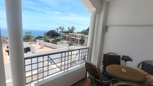 Un balcon avec vue sur les immeubles et la mer. On y trouve une table avec des chaises et un vase rempli de fleurs.