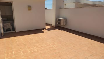 A rooftop terrace with a tiled floor, a white wall, and a door leading to a kitchen area.