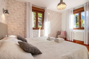 Un dormitorio con una cama grande, una mesita de noche, una lámpara y vistas al exterior a través de las ventanas.