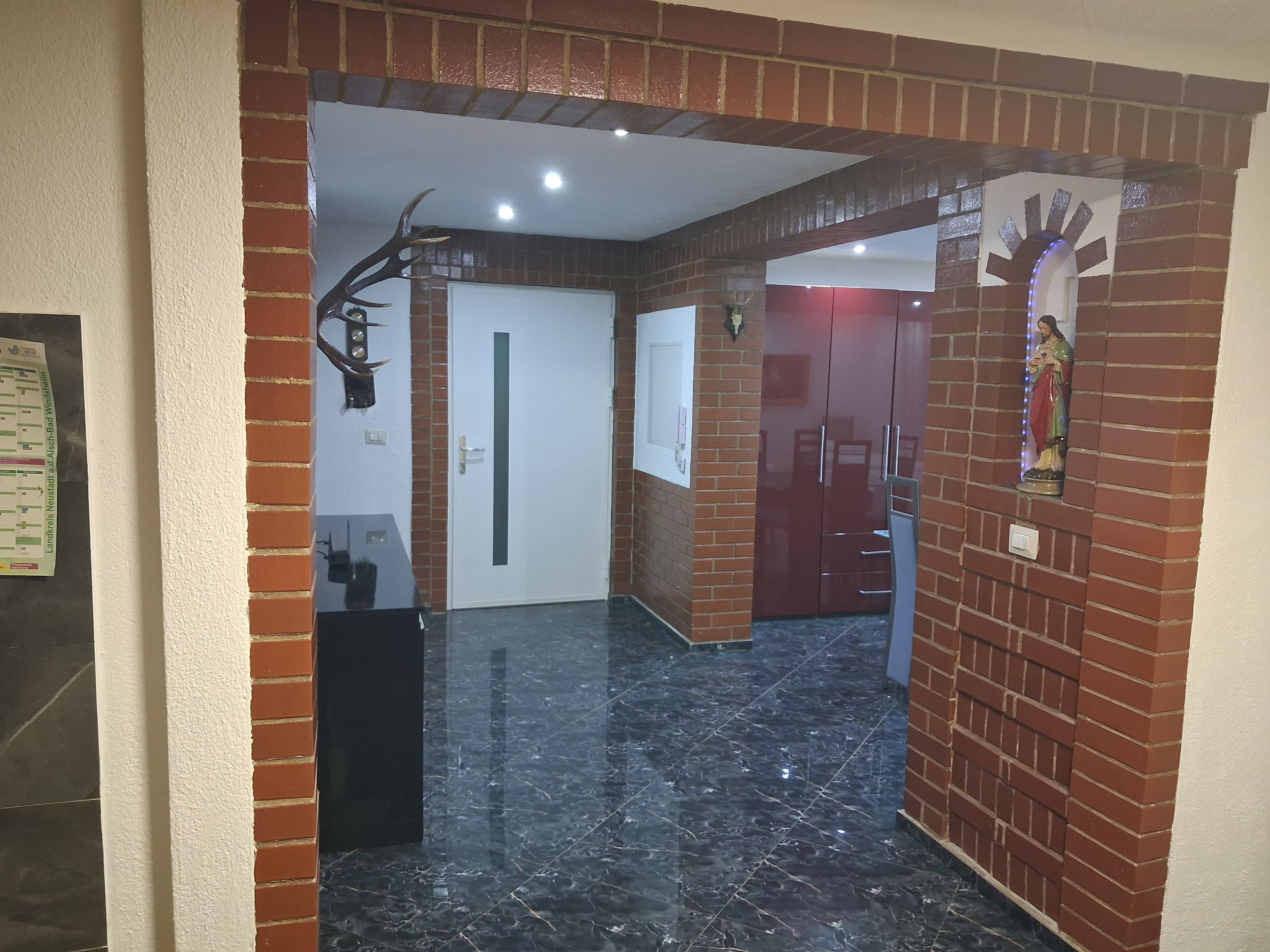 Un couloir avec une arche en briques, un sol en marbre noir, une porte blanche et une statue accrochée au mur.