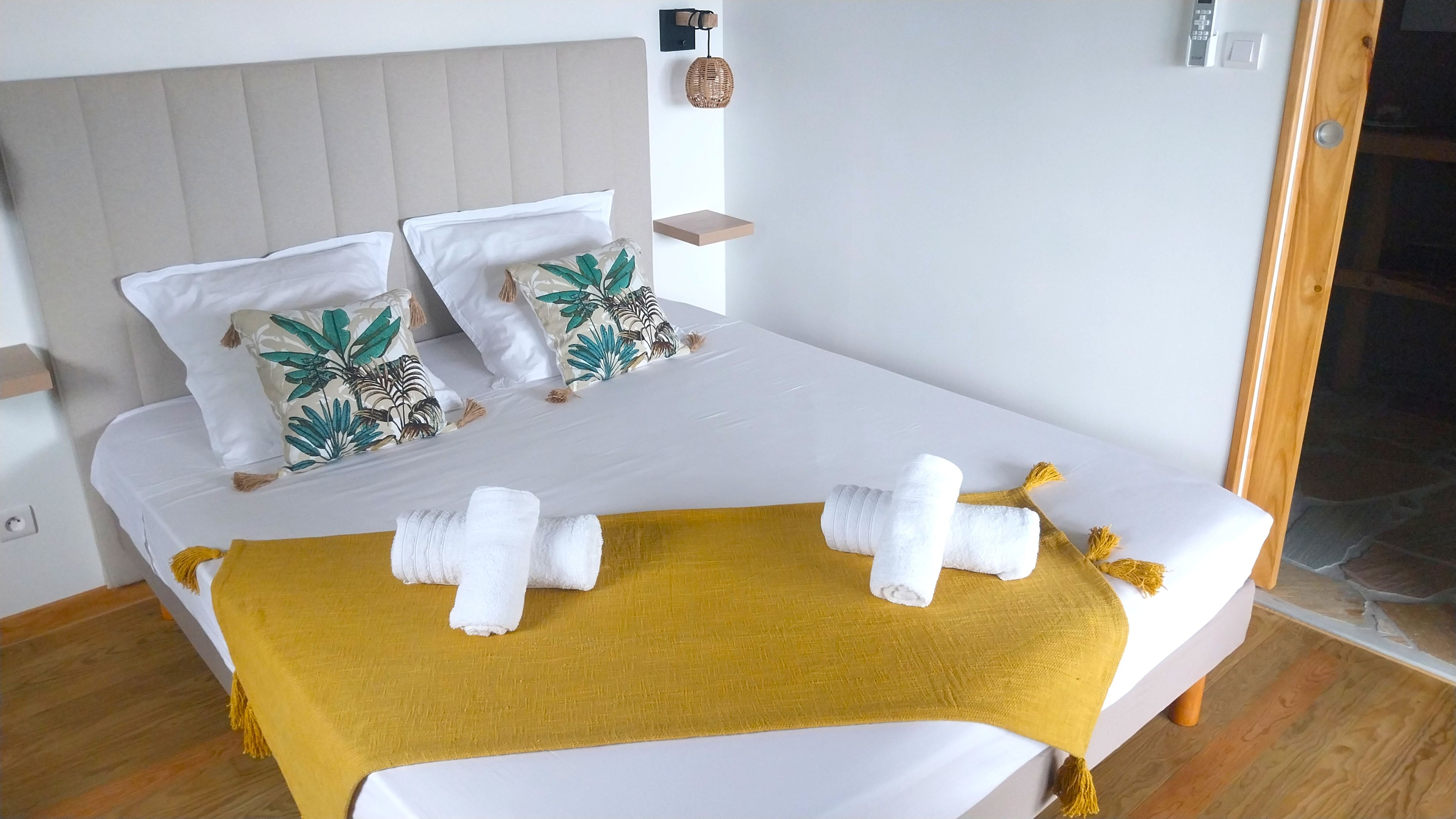 Un lit avec une literie blanche, deux coussins décoratifs, un plaid jaune et des serviettes enroulées.