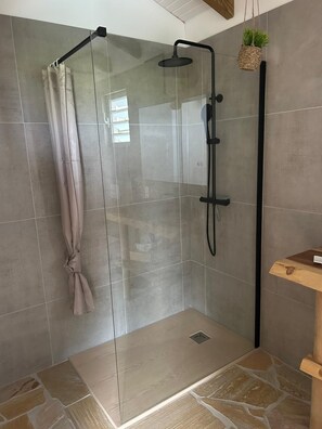 A glass-enclosed shower with a towel rack, a wooden shelf with a plant, and stone-patterned flooring.