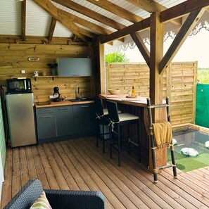 A wooden deck with a kitchen area, including a refrigerator, microwave, and a dining table with chairs.