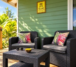 A wicker sofa set with colorful cushions on a wooden deck, a sign above reading 'RHUM BOGNE 5cl', and a wooden railing in the background.