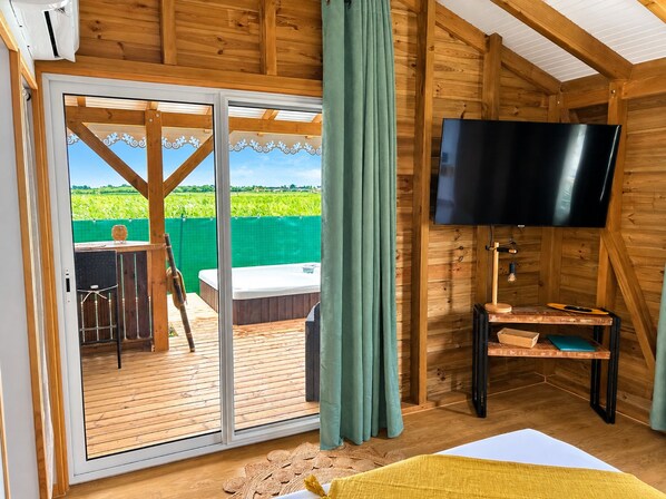 A wooden cabin with a sliding glass door leading to an outdoor deck, a flat-screen TV mounted on the wall, and a hot tub.