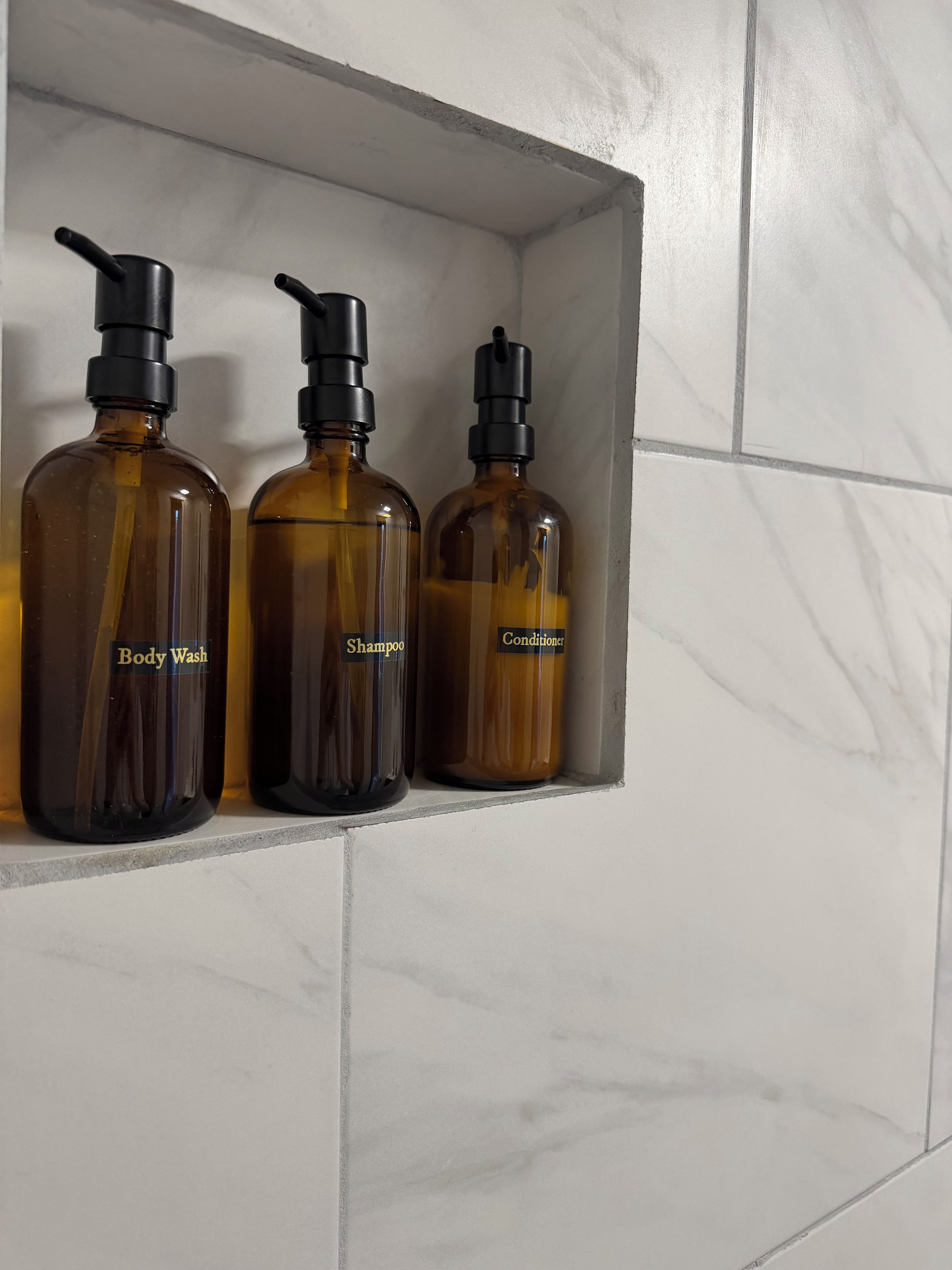 Three bottles of body wash, shampoo, and conditioner on a shelf against a tiled wall.
