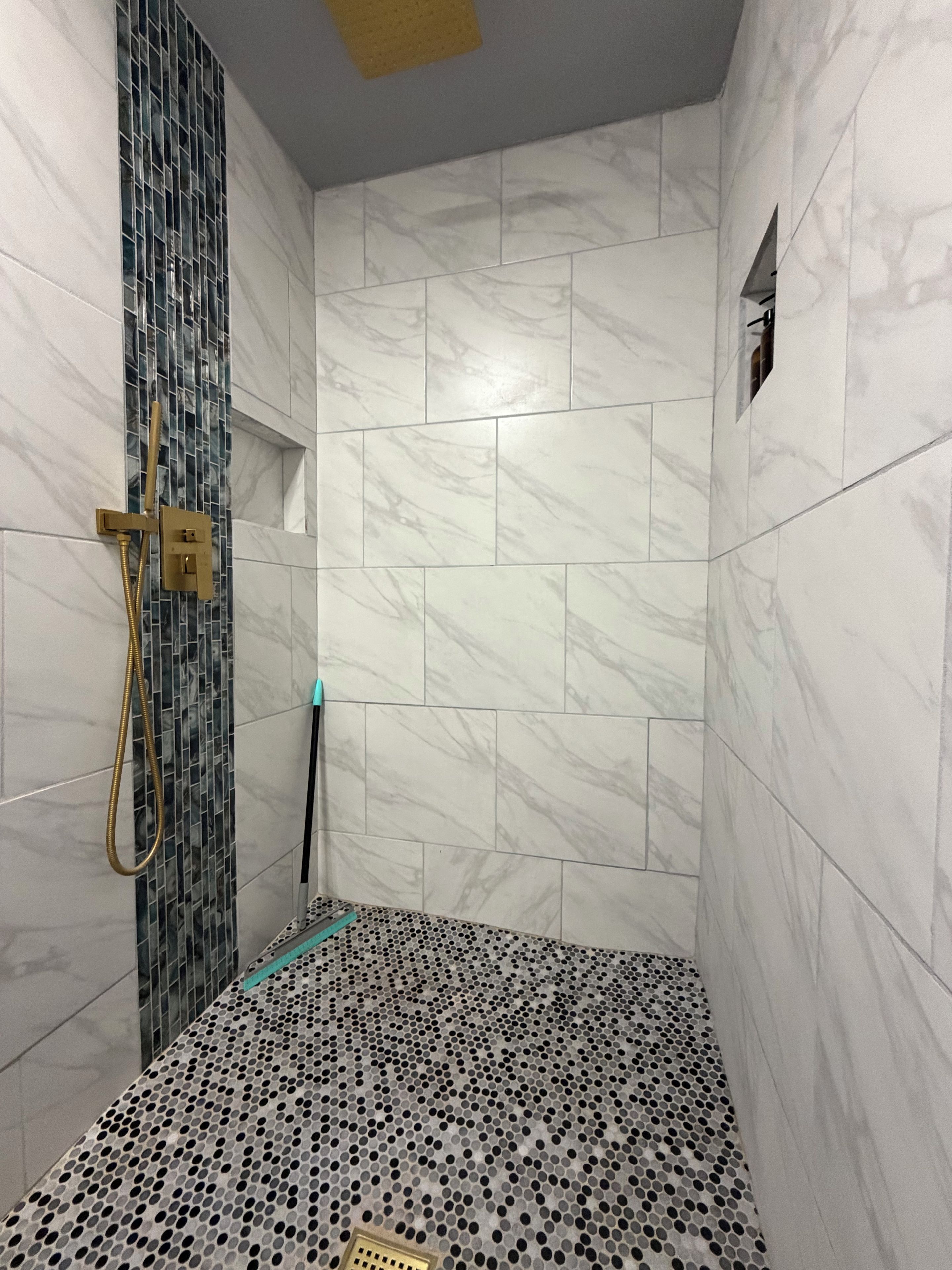 A bathroom with a shower area featuring a tiled floor with a perforated pattern, a showerhead, and a wall-mounted control panel.