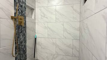A bathroom with a shower area featuring a tiled floor with a perforated pattern, a showerhead, and a wall-mounted control panel.