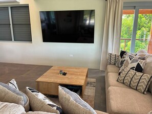 Ein Wohnzimmer mit einem an der Wand montierten Flachbildfernseher, einem Holztisch und einem beigefarbenen Sofa mit gemusterten Kissen.