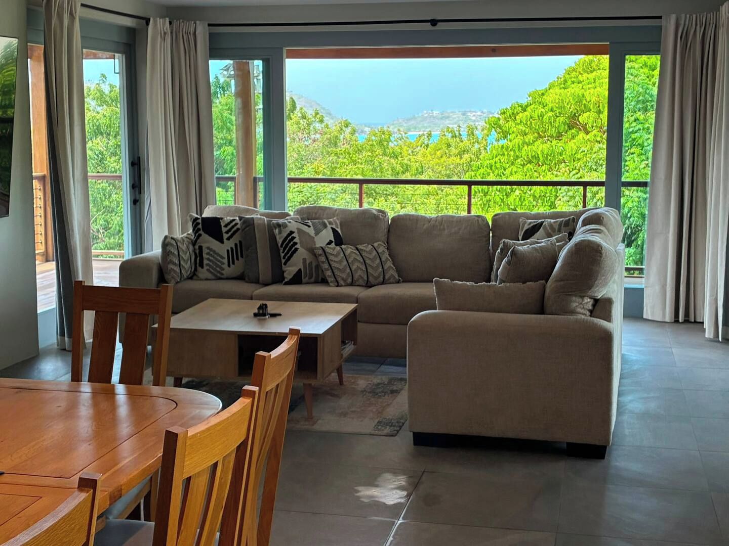 Ein modernes Wohnzimmer mit einer Couch, einem Holzesstisch und Stühlen. Große Fenster bieten Blick auf Grünflächen und Hügel.