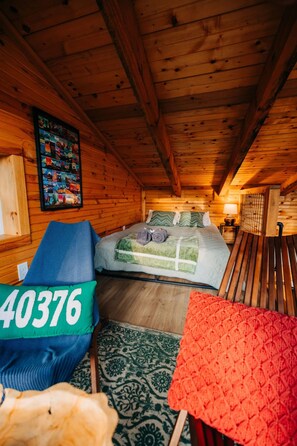 A cozy cabin room with a bed, a blue chair, a green pillow with numbers, a red blanket, and a wooden ceiling. - Tunnelvision Tiny Cabin Red River Gorge, KY Hot Tub Wifi Pet Friendly Private (Stanton)