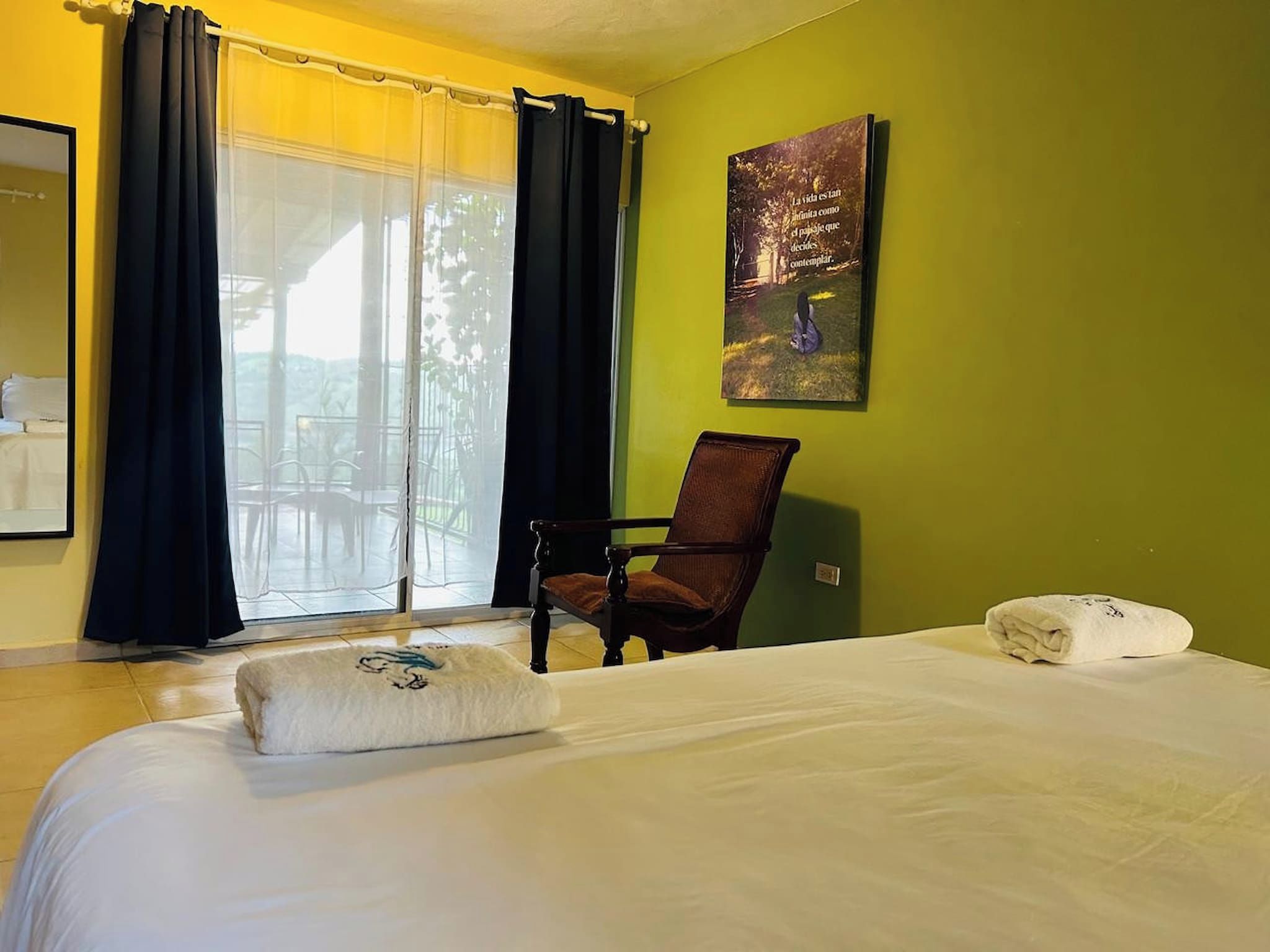 Une chambre d’hôtel avec un lit, une chaise, une fenêtre avec des rideaux et un tableau au mur.