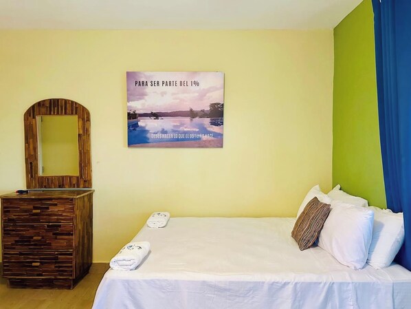 A hotel room with a bed, a wooden dresser, a mirror, and a poster on the wall.