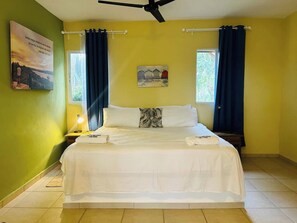 A hotel room with a bed, two windows with curtains, a painting on the wall, and a ceiling fan.