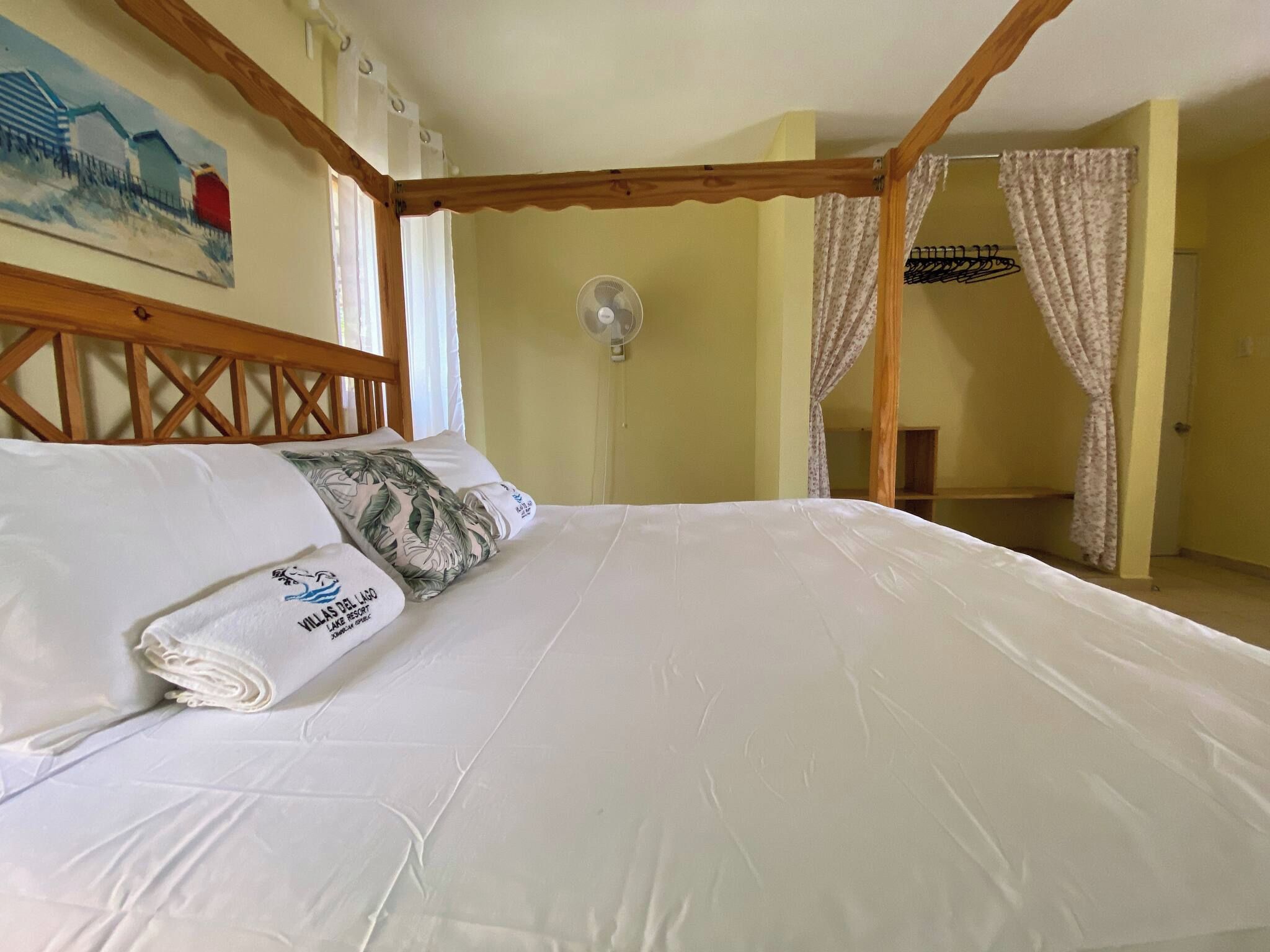 A bedroom with a wooden headboard, a bed with white linens, a fan, and a painting on the wall.