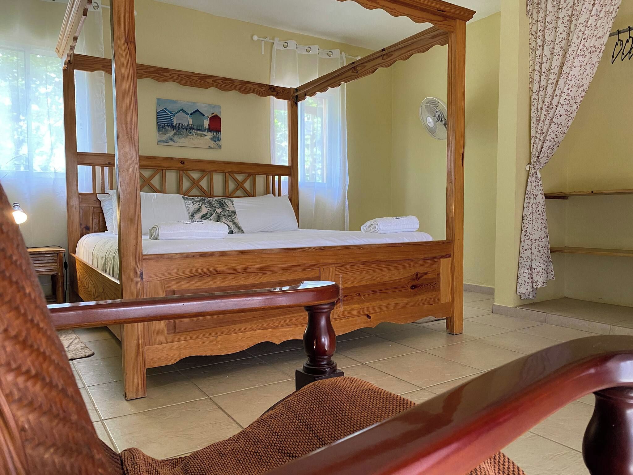 Een hemelbed met houten frame, een schilderij aan de muur en een ventilator.