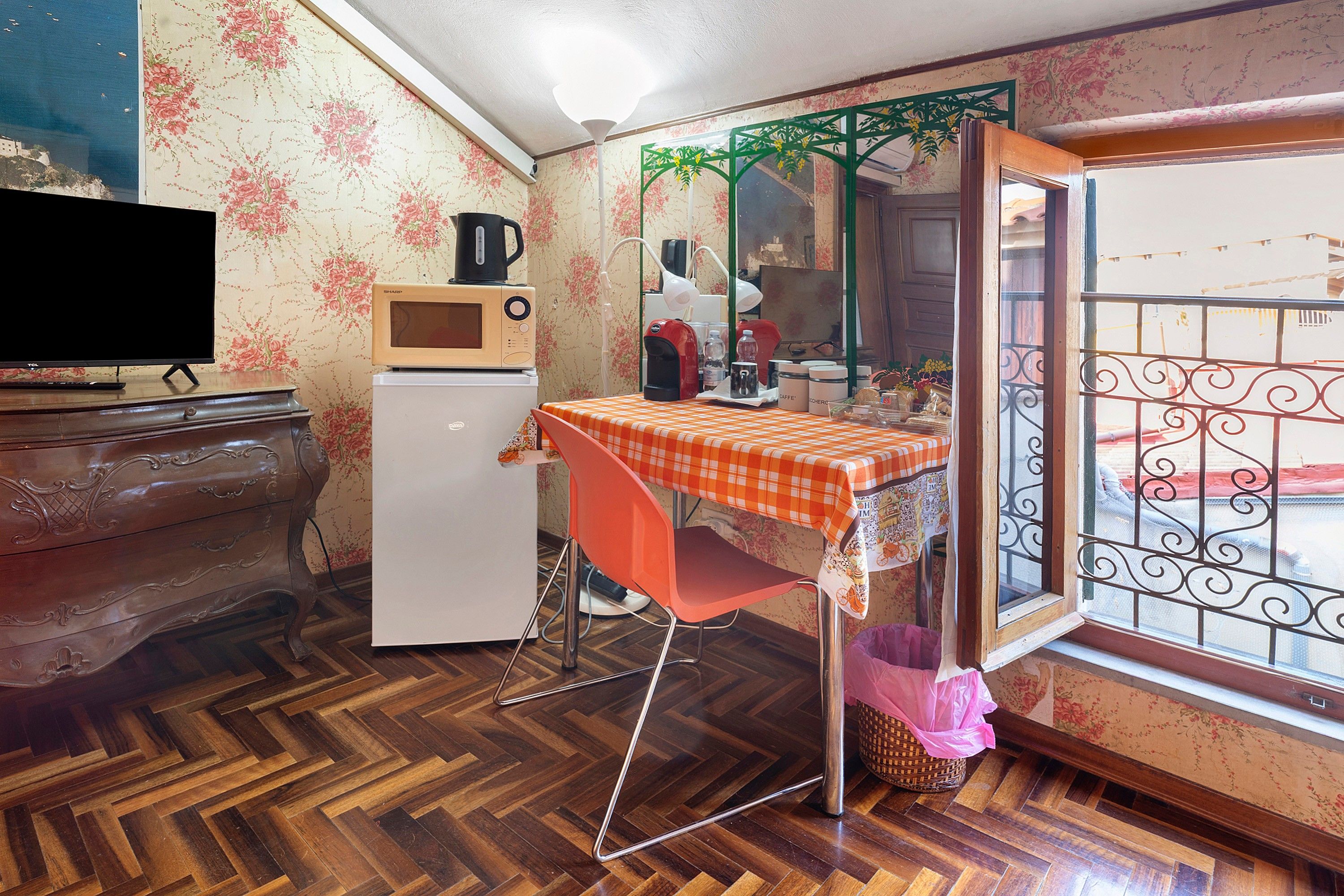 Una stanza compatta con pavimento in parquet a spina di pesce, una televisione, un forno a microonde, un frigorifero, un tavolo con tovaglia a quadretti, una sedia rossa e una finestra con inferriate decorative.