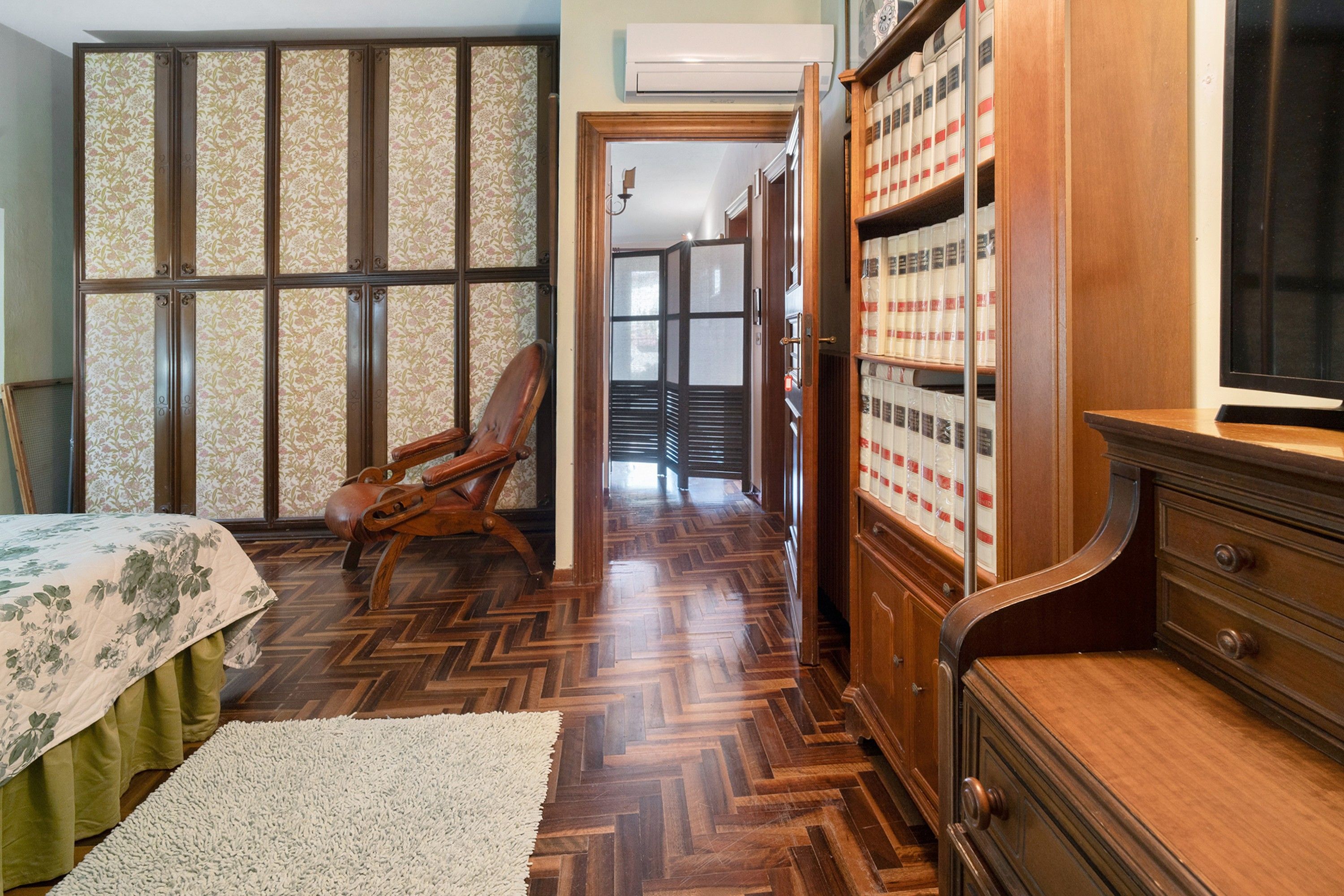 Een kamer met een visgraatparketvloer, een houten stoel, een bed met een bloemenprintdeken, een houten dressoir en een boekenkast met boeken.