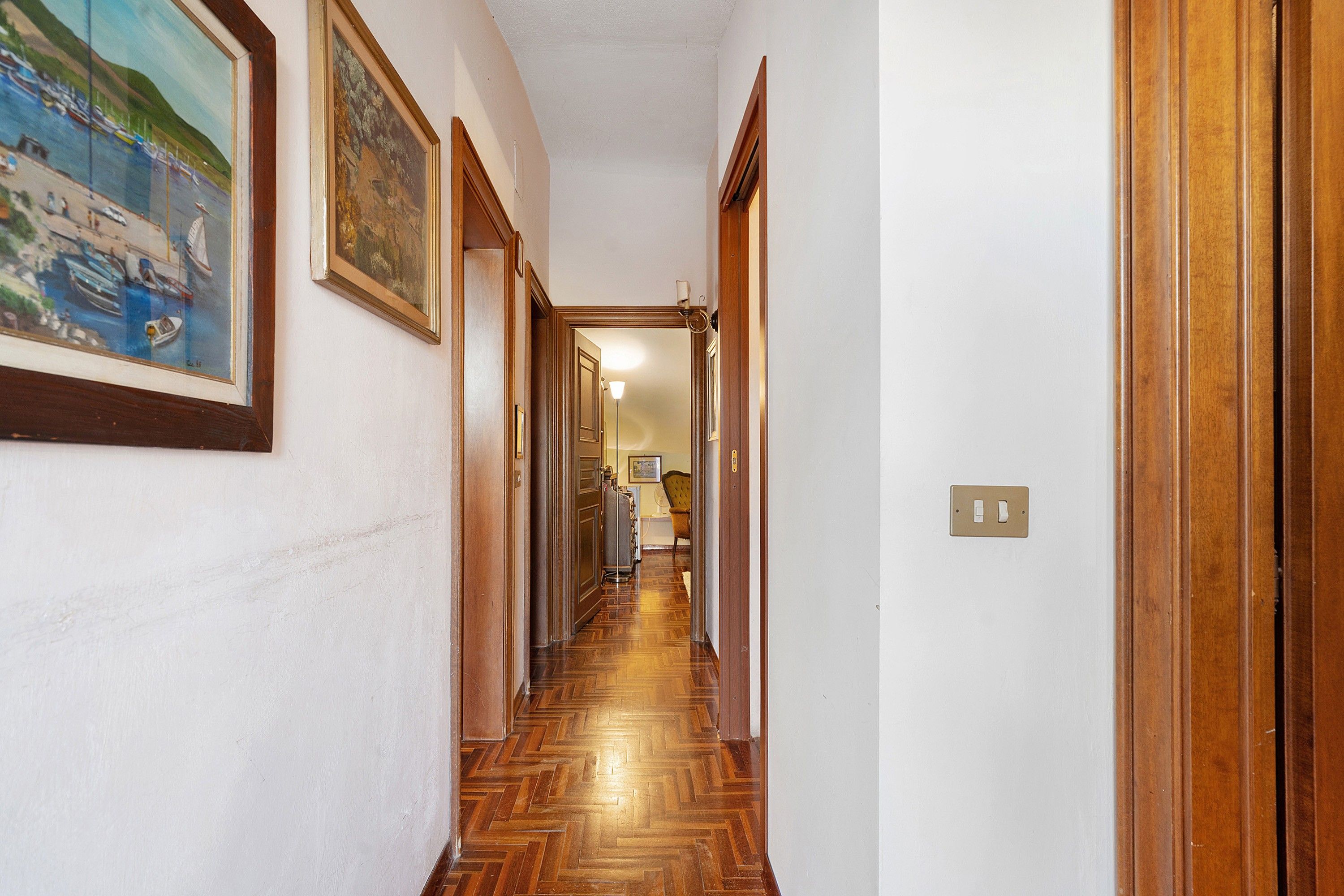A hallway with wooden flooring, framed artwork on the walls, and a door leading to another room.