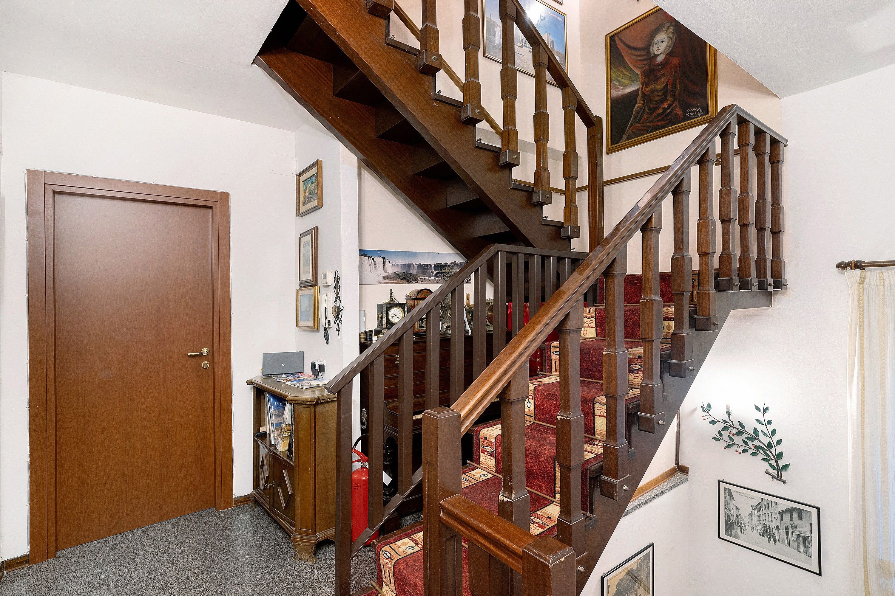 A wooden staircase with a red carpeted landing, a closed wooden door, and a wall with framed pictures and a clock.