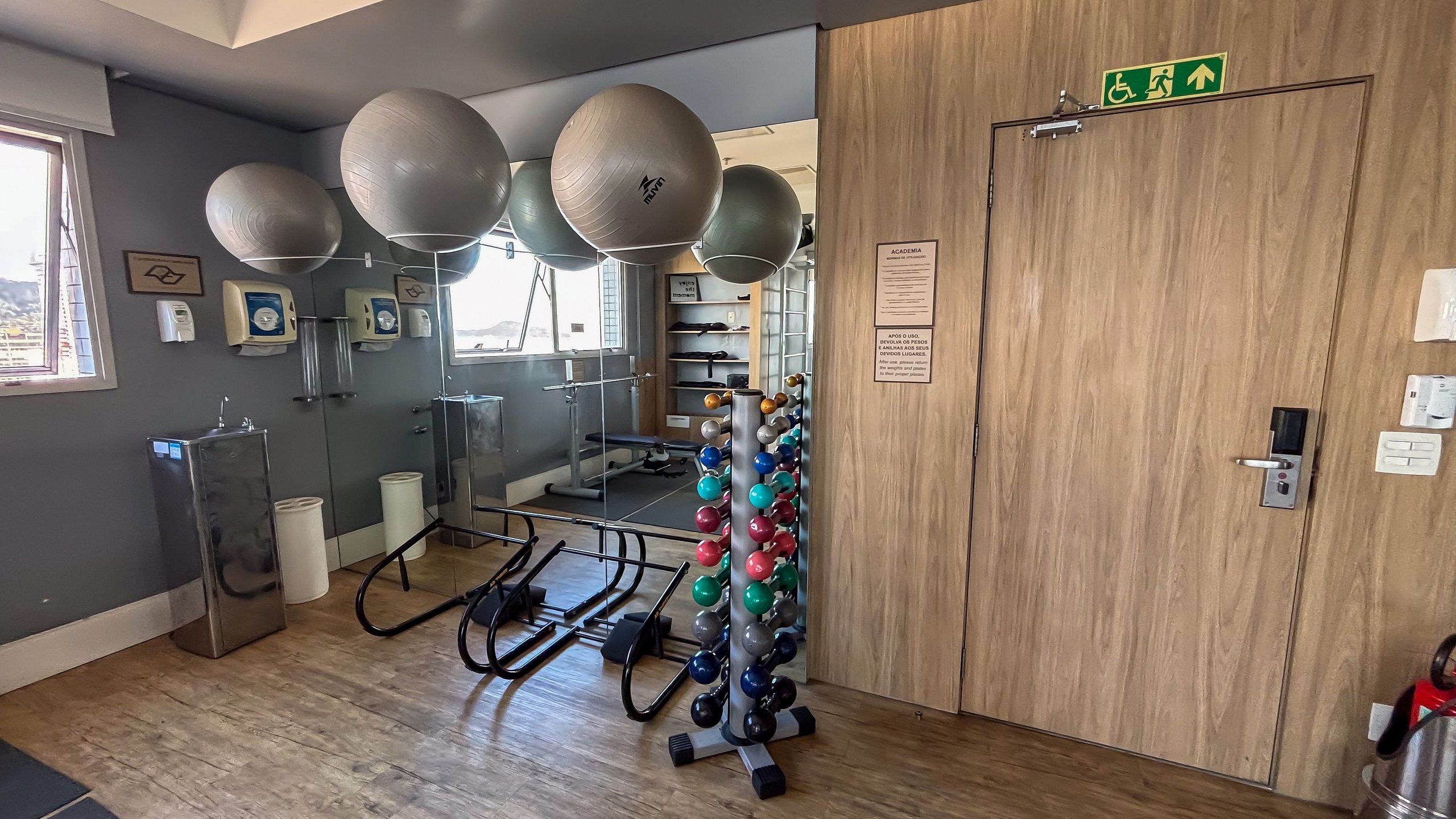 A gym with exercise equipment, a water dispenser, and a wooden door with an emergency exit sign.