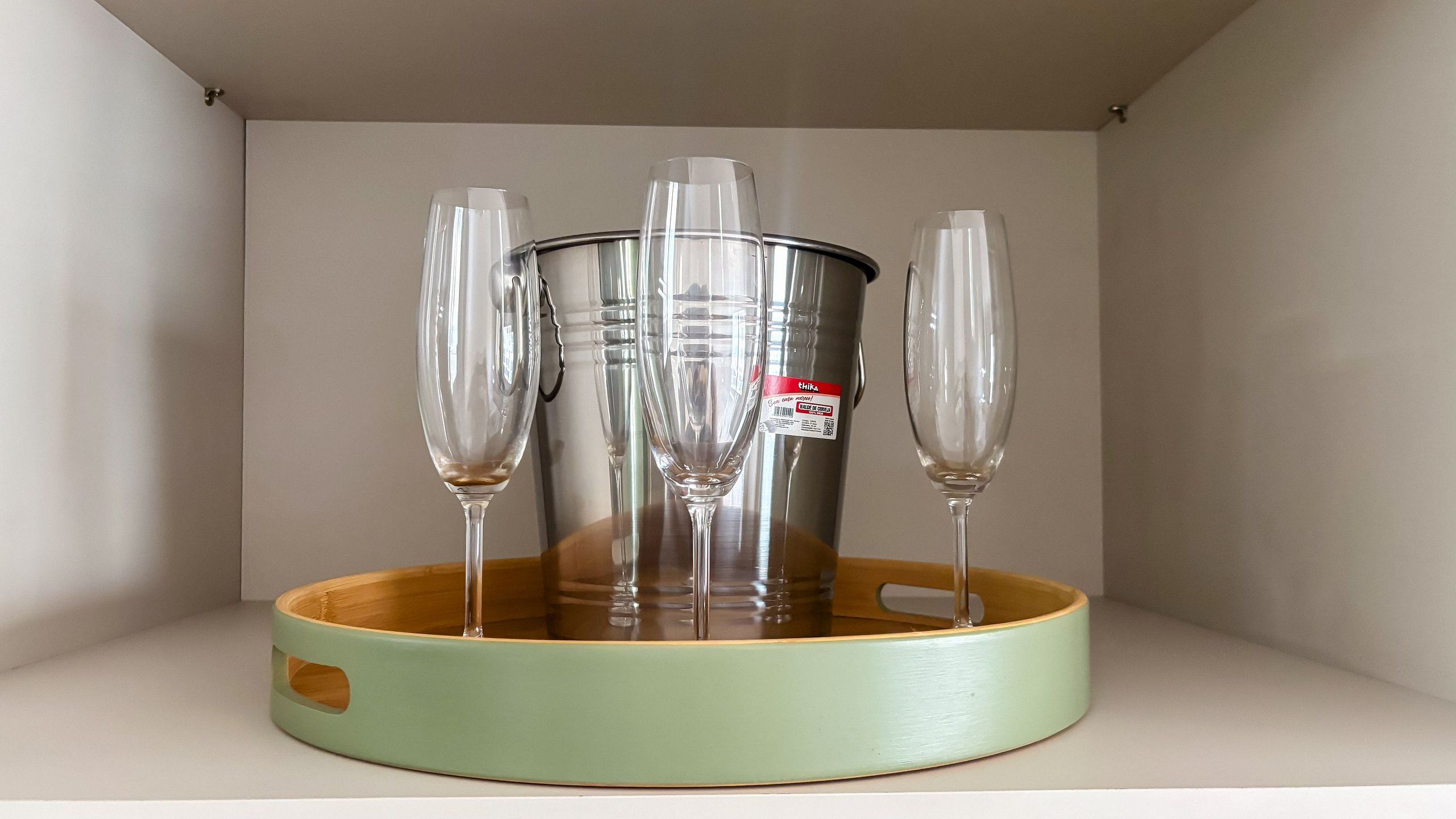 A metal bucket with a handle and a label, surrounded by four clear glasses on a wooden tray.